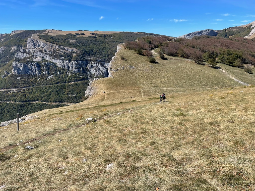 a cyclist pushing a bike up a steep grassy slope, with limestone cliffs and autumn forests in the background