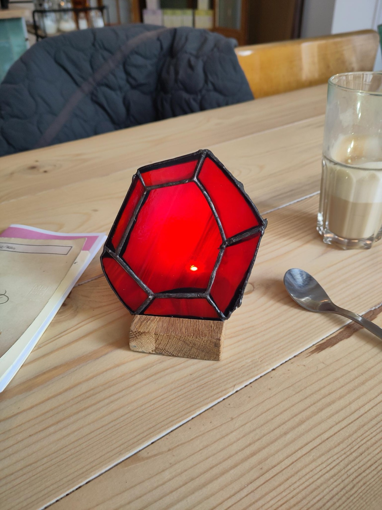 the freeform red ruby stained-glass panel standing on a small oak block on a wooden table, glowing from a flashlight placed behind it