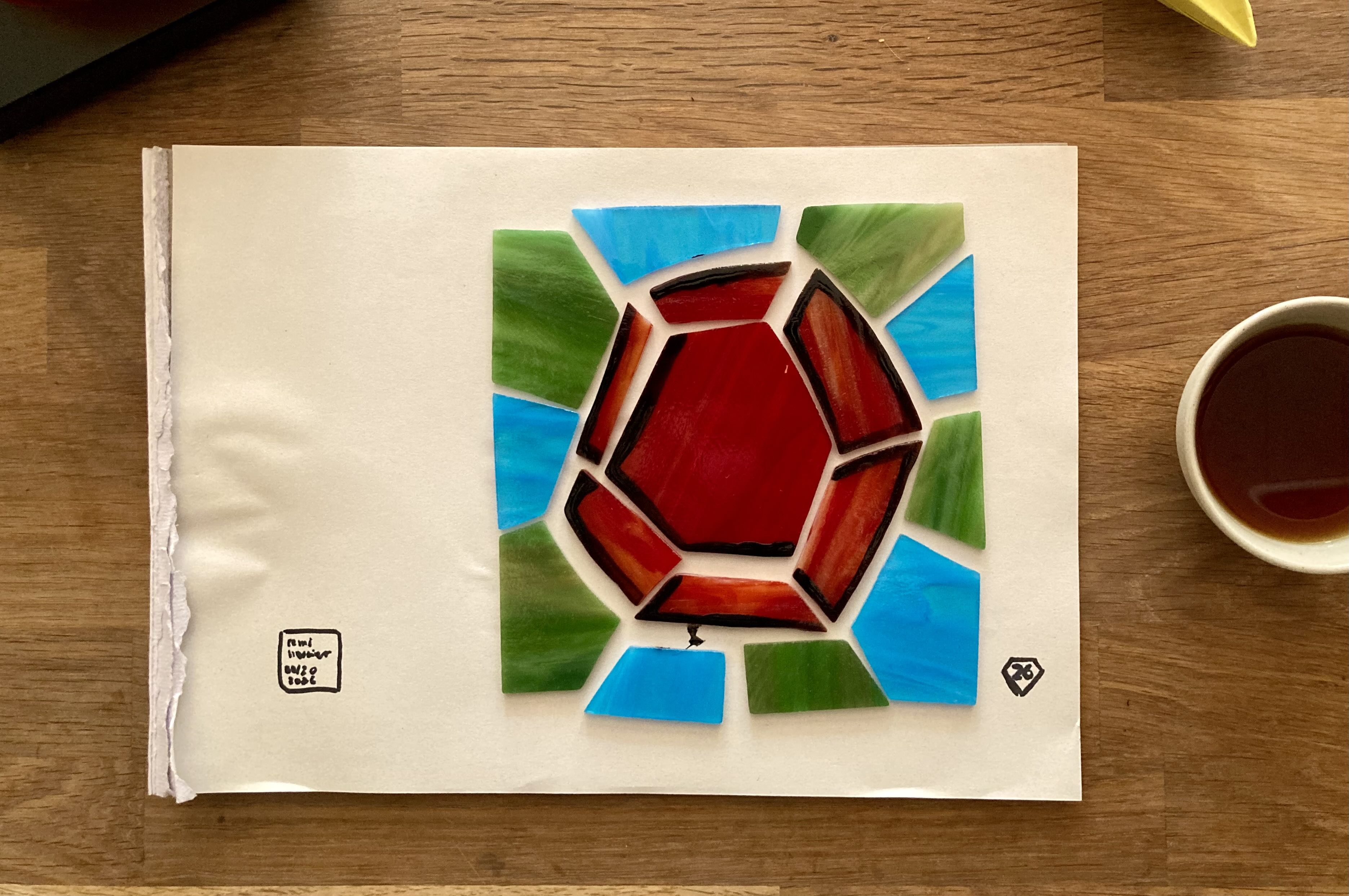 the pieces of the square stained-glass panel laid out on paper: a red ruby surrounded by alternating light blue and green glass pieces