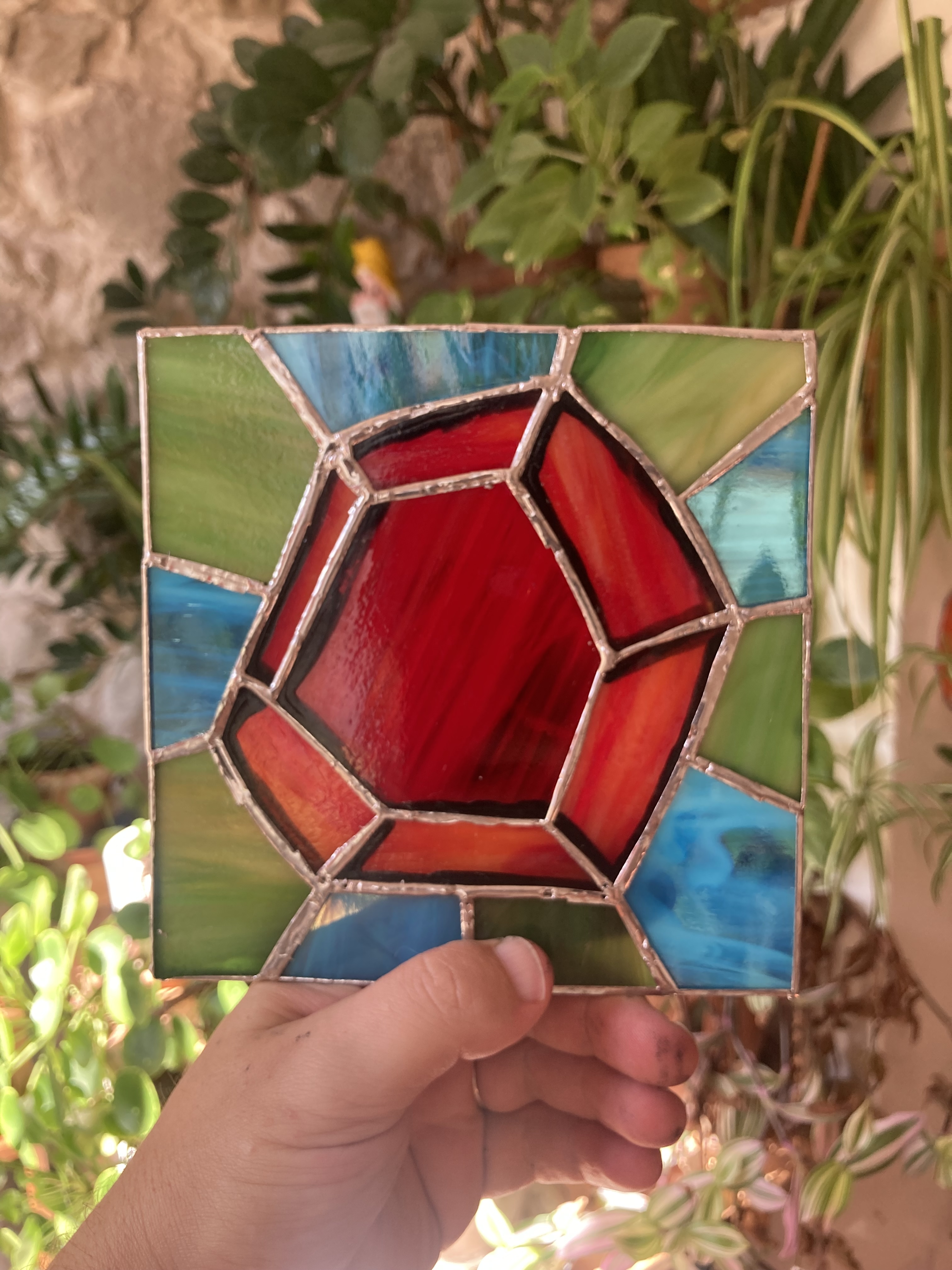 a hand holding the finished square stained-glass panel, a red ruby surrounded by light blue and green glass, in front of houseplants