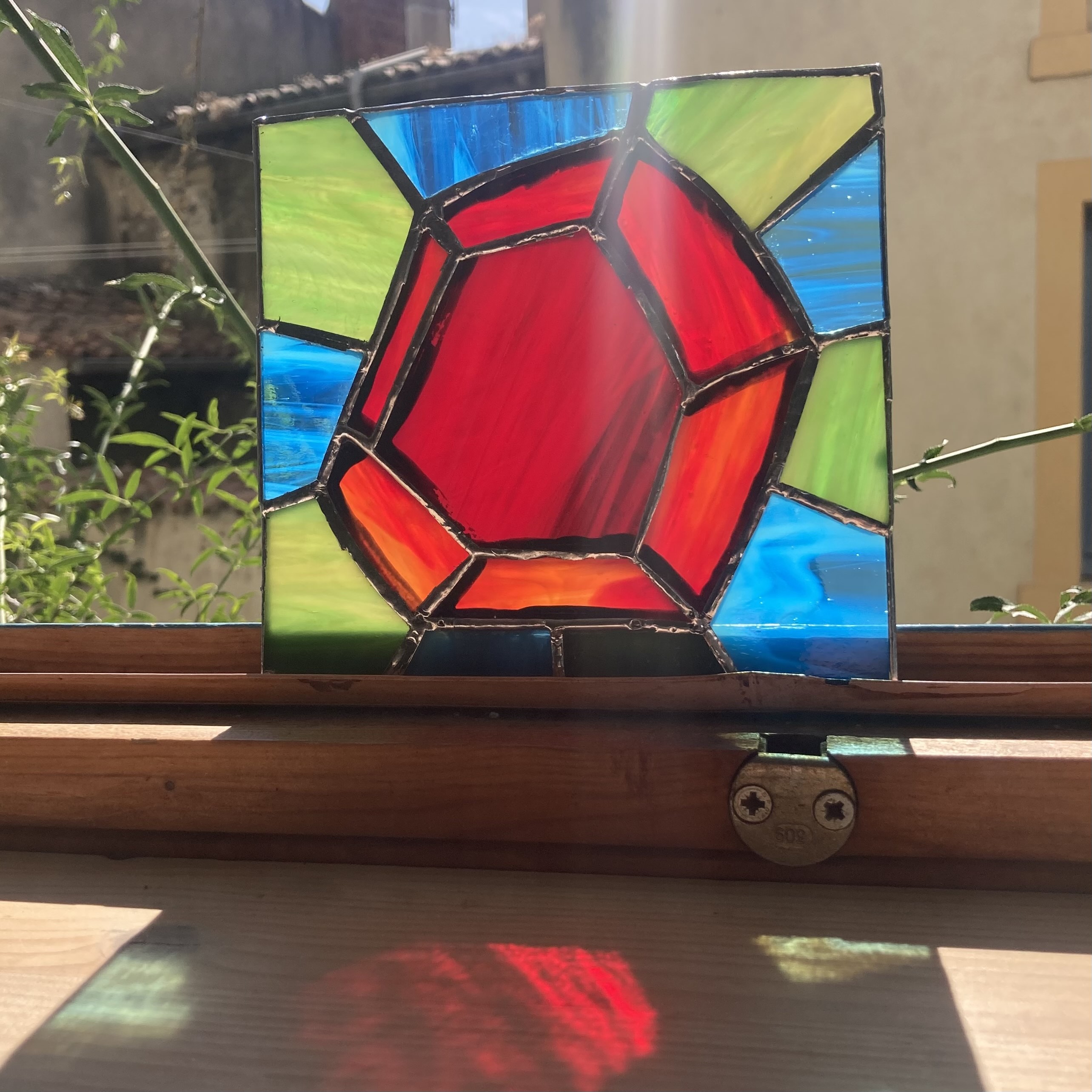 the finished square stained-glass panel standing on a sunny windowsill, casting red light onto the wooden sill